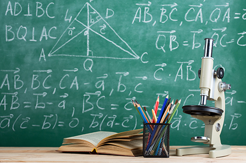 A book, microscope and pot of pencils on a desk infront of a chalk board with physics written on.