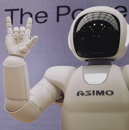 White robot with a black screen face doing the 'i love you' gesture in American Sign Language